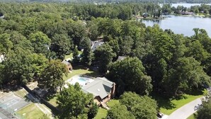 An aerial view of a residential area with a swimming pool, a tennis court, and a large body of water in the background. - Lake living in a relaxed family and pet-friendly home (Chapin)