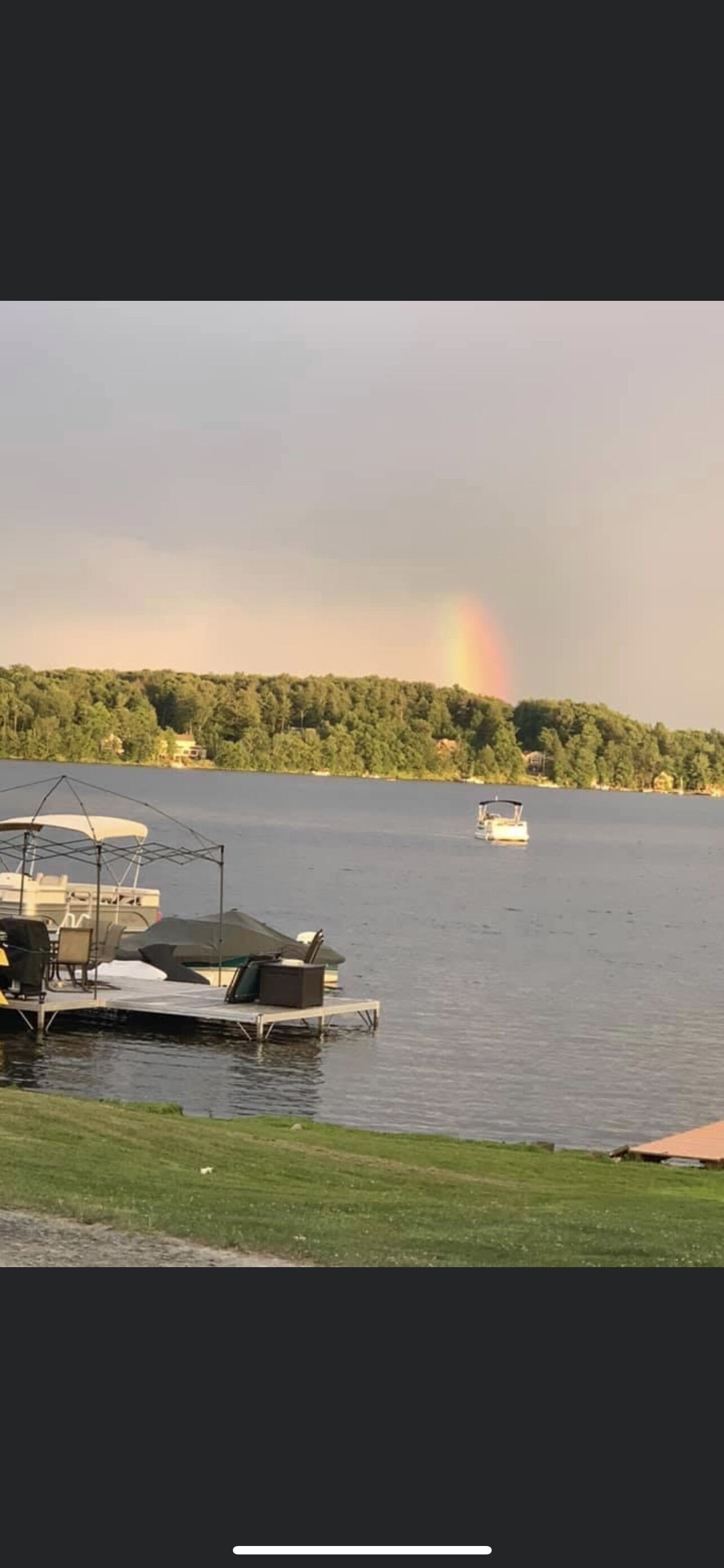 Une vue sur un lac, avec un quai, un bateau et un arc-en-ciel au loin.