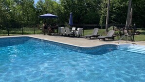 Une piscine entourée d’une clôture et d’arbres, avec des chaises longues et des parasols.