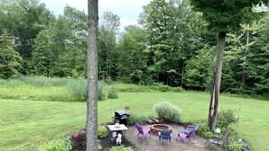 Une terrasse aménagée avec du mobilier d’extérieur, un barbecue et un foyer, le tout entouré de parterres et d’arbres.