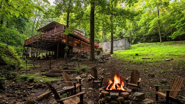 Una cabaña con terraza, rodeada de un bosque. Hay un brasero con sillas a su alrededor.