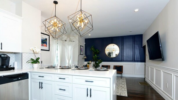 Une cuisine moderne avec des meubles blancs, un îlot central, des luminaires suspendus et un mur d’accent bleu foncé.