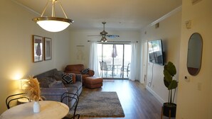 A spacious living room with a sofa, ottoman, and a dining table. There is a ceiling fan, a TV mounted on the wall, and a large window with curtains.