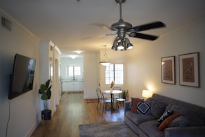 A living room with a gray sofa, a coffee table, and a dining area with a table and chairs. There is a ceiling fan, a TV mounted on the wall, and a plant in the corner.