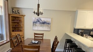 A dining area with a wooden table and chairs, a chandelier, a bookshelf with decorative items, and a painting on the wall. - Townhouse backing onto the skating loop (Apex Mountain, Penticton)