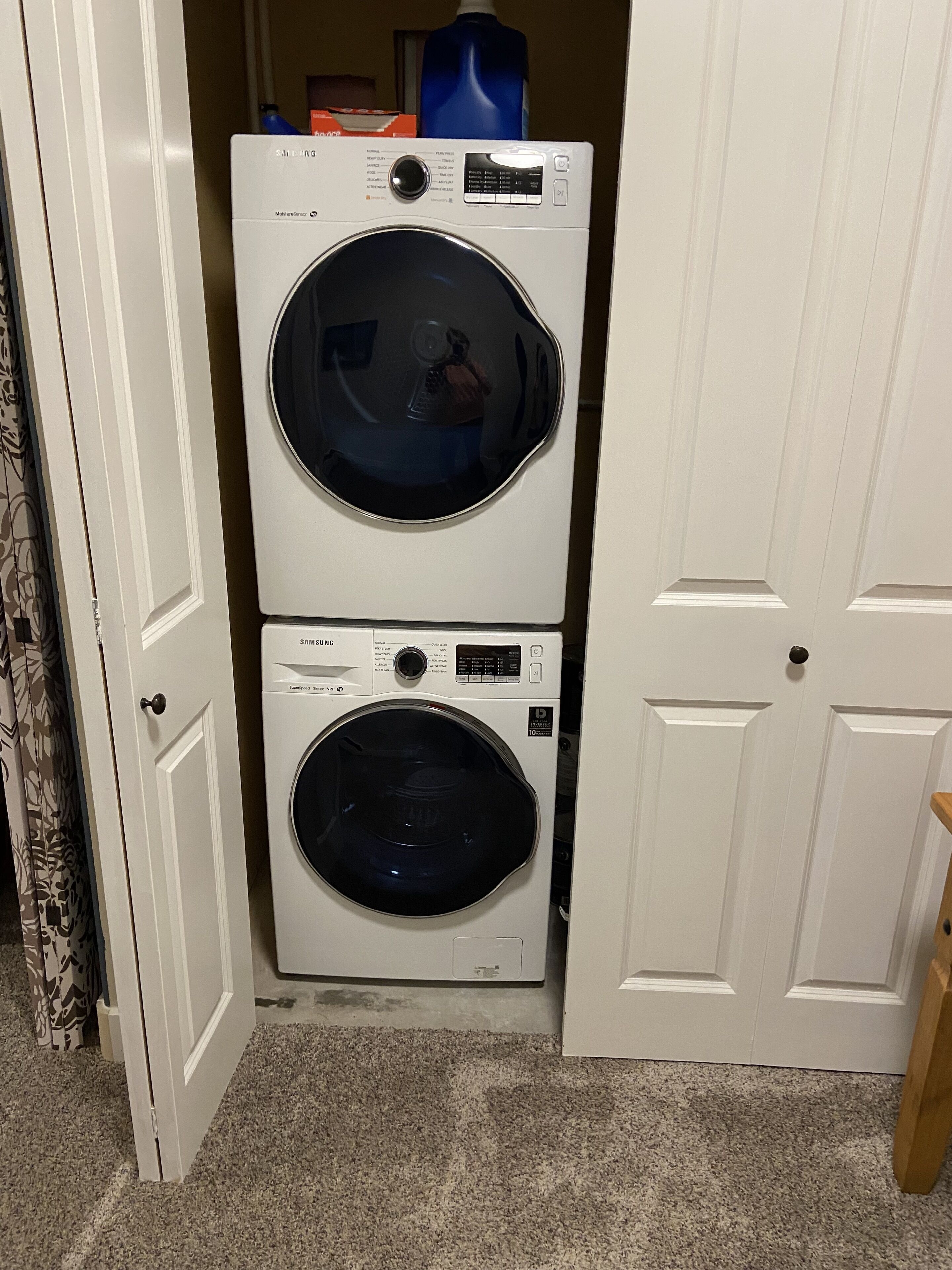 A laundry room with two stacked washers and dryers, a blue detergent bottle, and a door leading to another room.