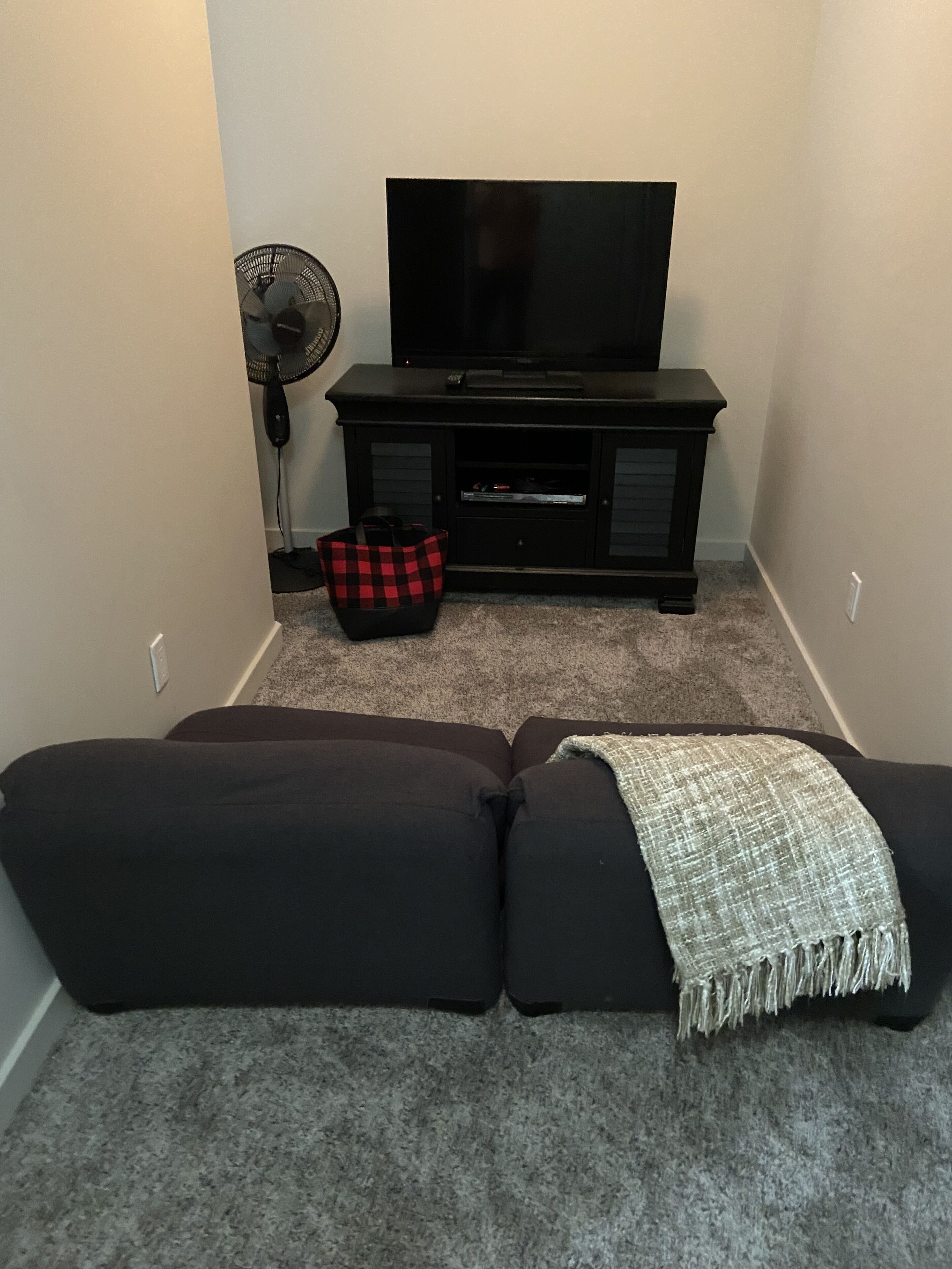 A living room with a black entertainment unit, a flat-screen TV, a red and black checkered bag, and a gray blanket on a black sofa.