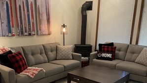 A living room with a sectional sofa, a coffee table, a fireplace, and a large window with blinds. - Townhouse backing onto the skating loop (Apex Mountain, Penticton)