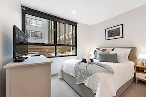Quarto com cama, mesa de cabeceira, abajur e janela com vista para a cidade.