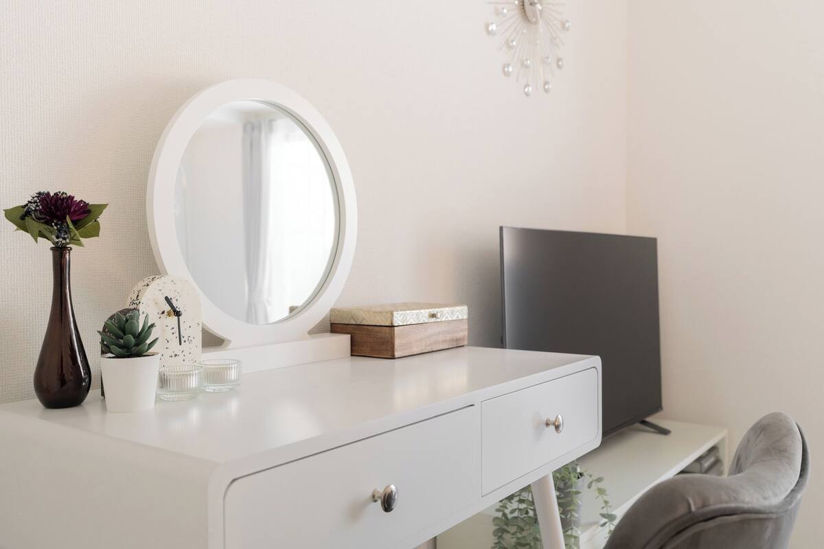 A white vanity with a round mirror, a dark vase with a plant, a small potted plant, and a box on the counter.