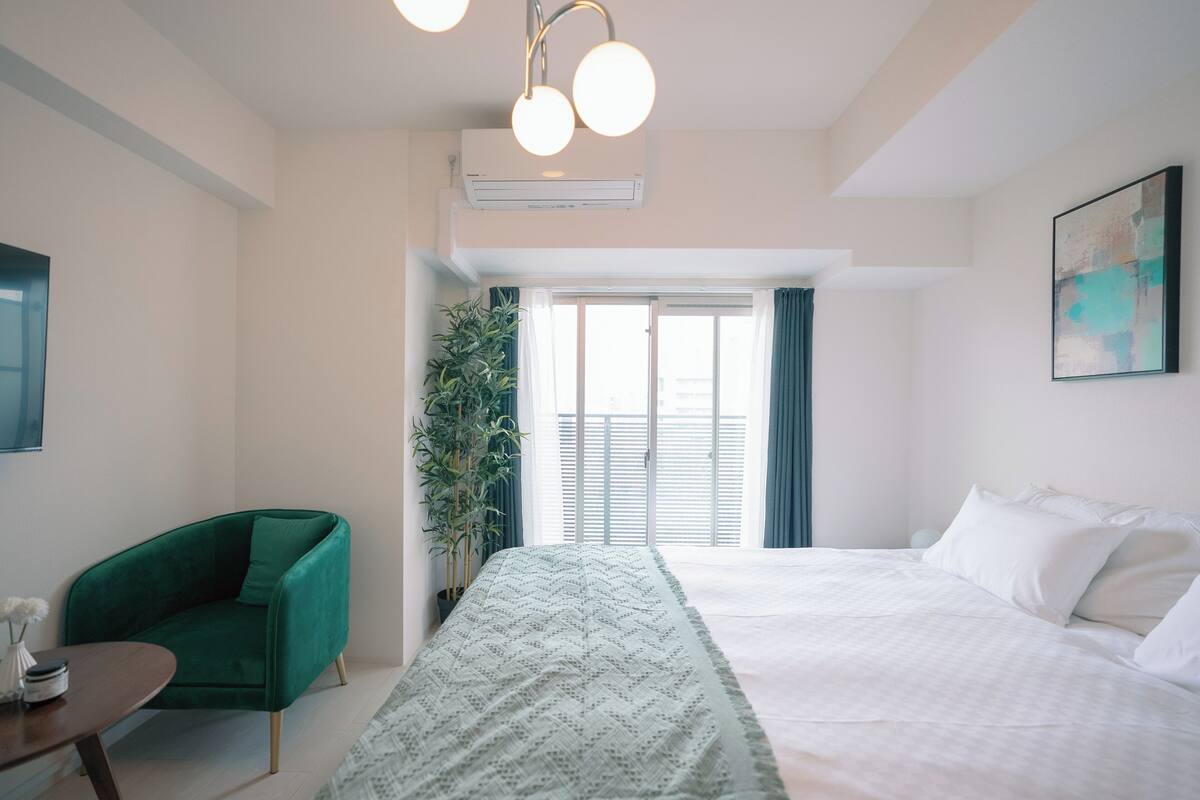 A modern hotel room with a large bed, a green armchair, a small table, and a potted plant.