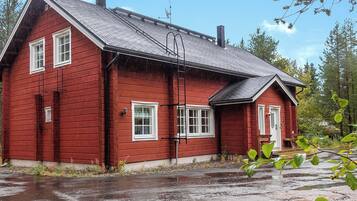 A red wooden house with a sloped roof, a chimney, and a small porch with a bench.