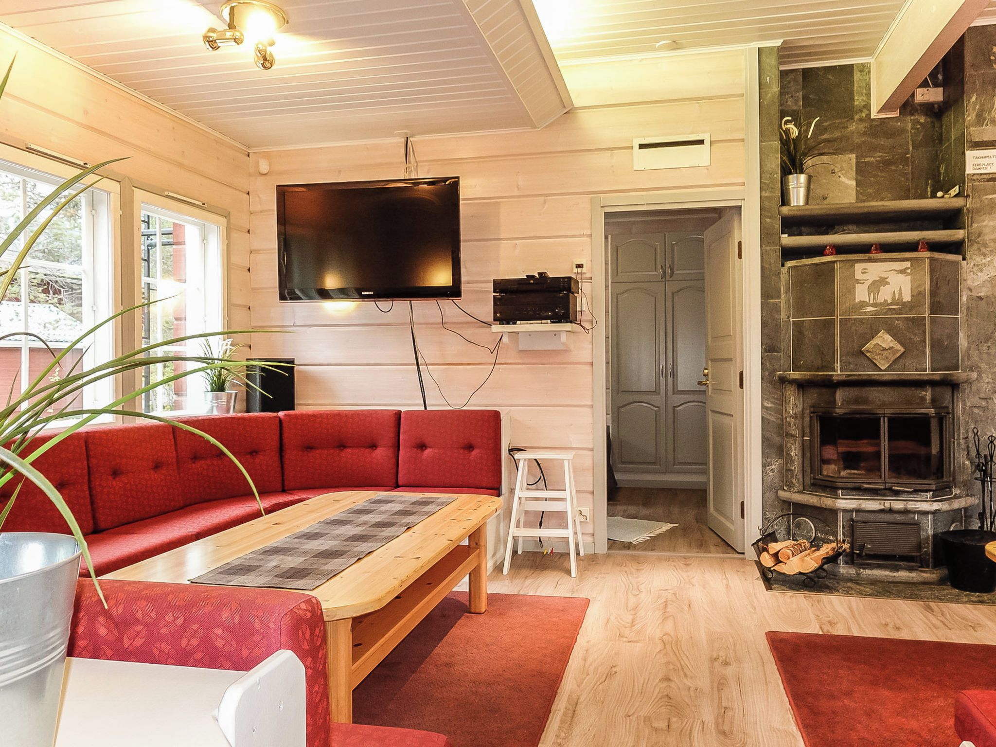 A living room with a red sofa, a wooden coffee table, a flat-screen TV mounted on the wall, a fireplace with a stone surround, and a door leading to another room.
