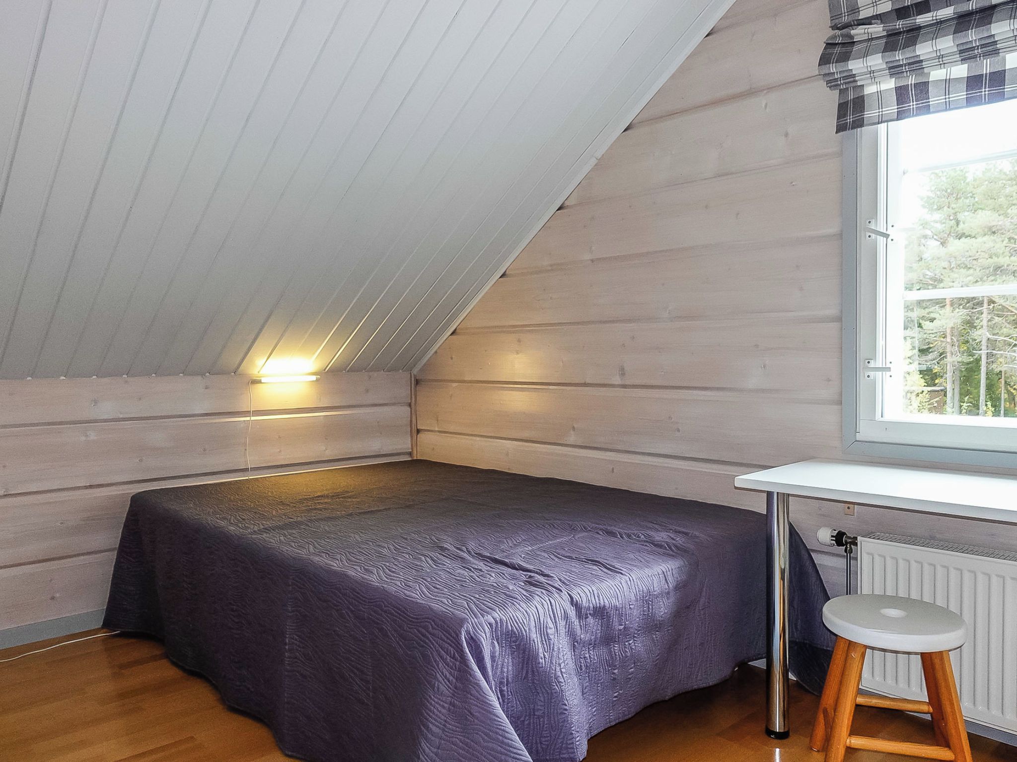 Una habitación pequeña con cama individual, techo inclinado, ventana con vistas a árboles y un radiador.