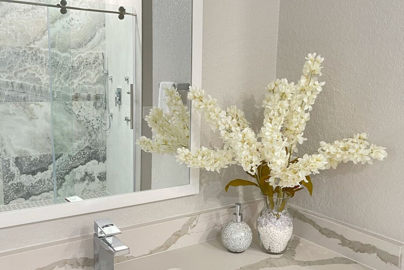 A bathroom with a glass shower enclosure, a white sink, and two decorative vases with white flowers.