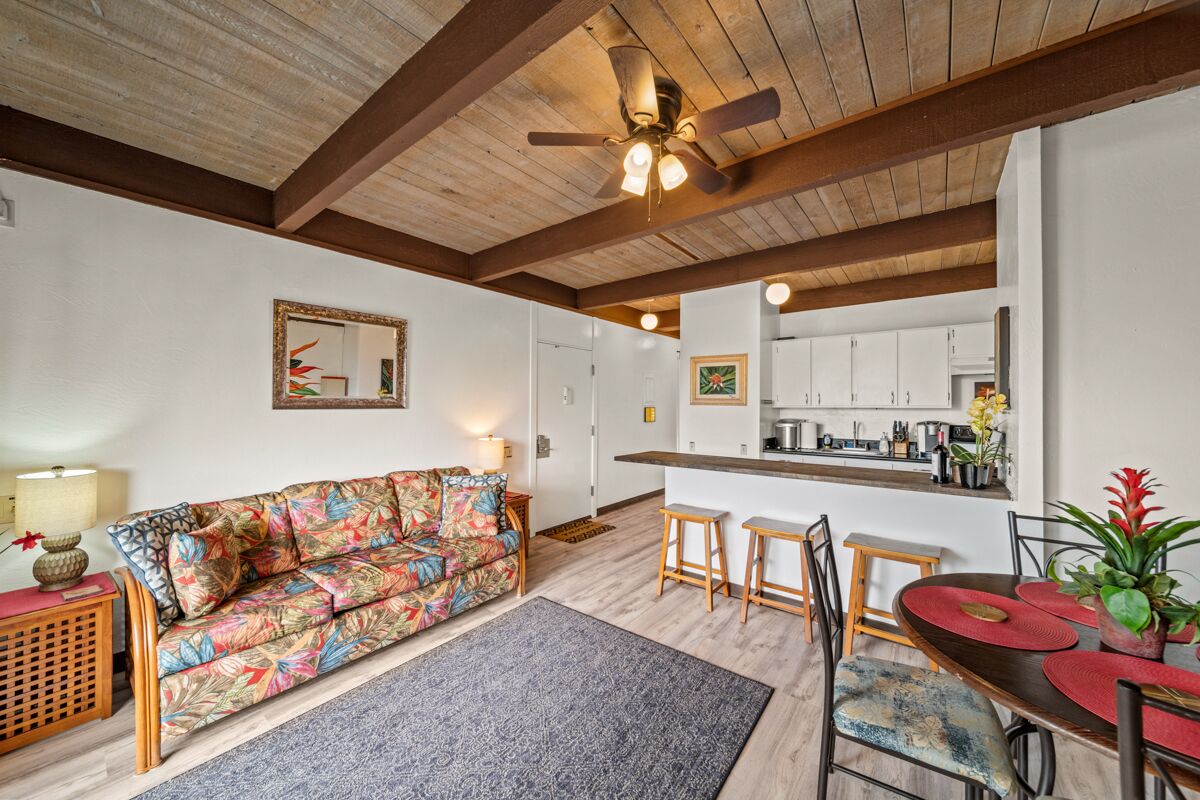 A living room with a sofa, a dining table with chairs, a kitchen area, and a ceiling fan.