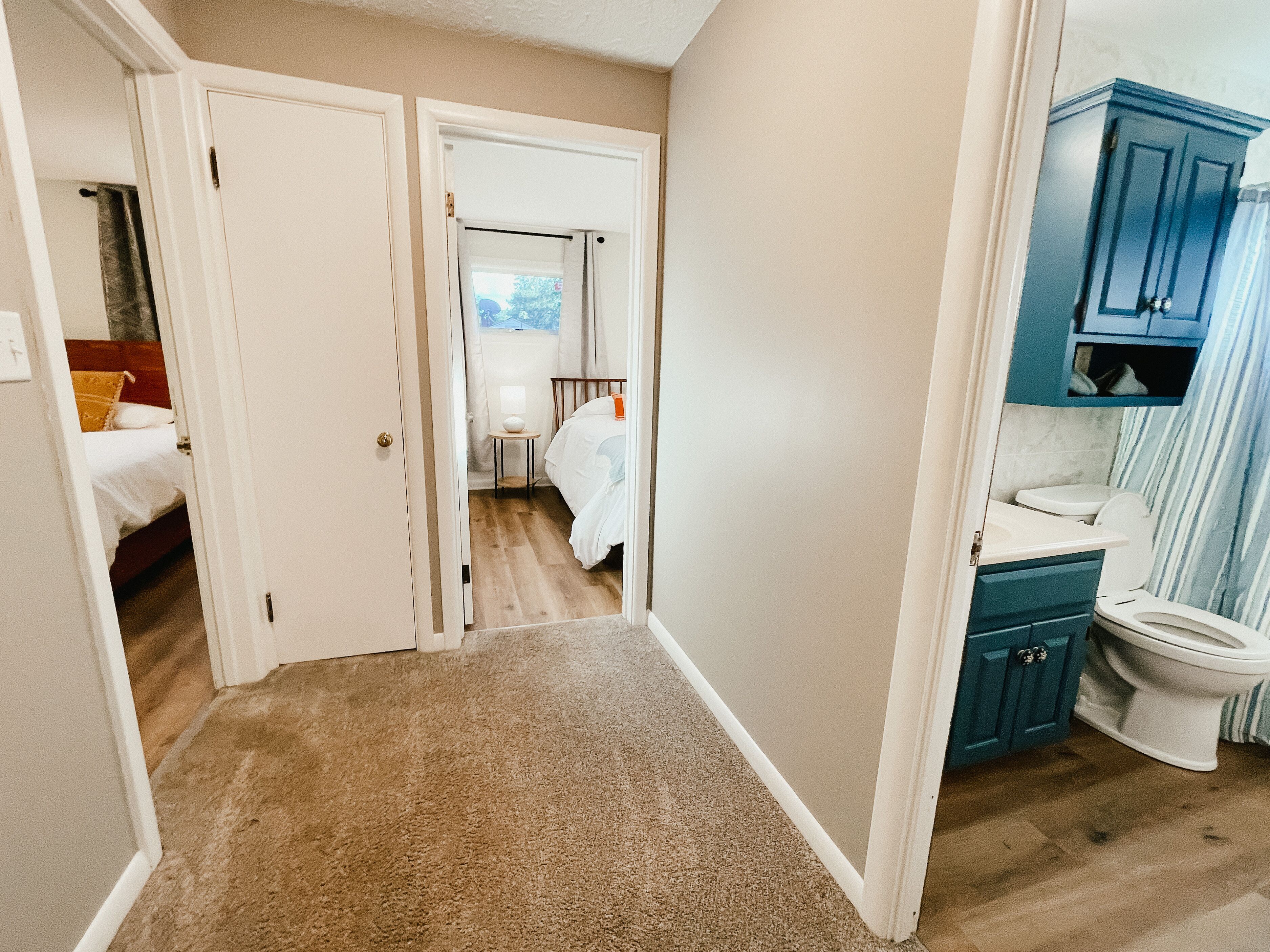 A hallway leading to a bedroom with a bed, a bathroom with a toilet and sink, and a small table with a lamp.