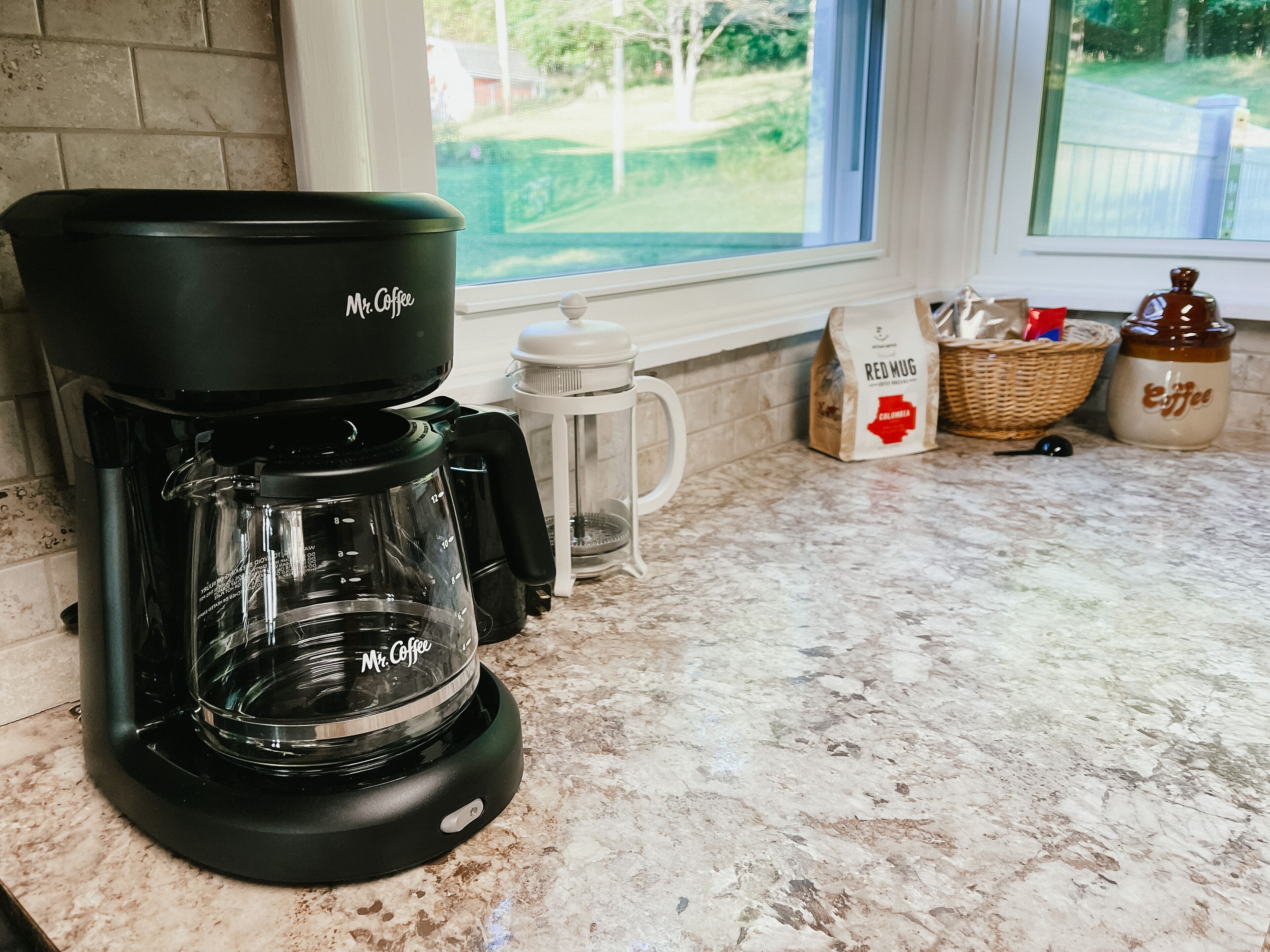 Une machine à café de la marque « Mr. Coffee » est posée sur un plan de travail de cuisine, près d’une fenêtre.