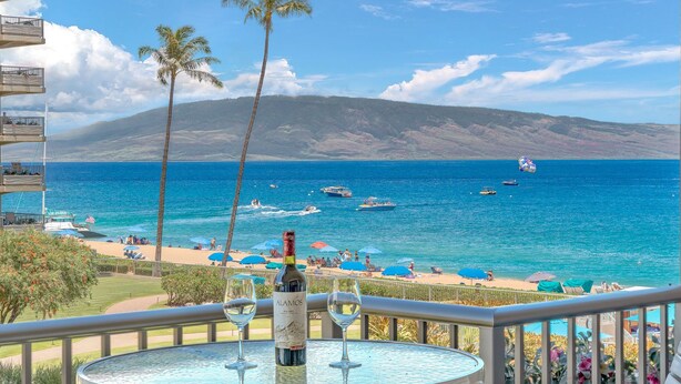A balcony with a table set for two, overlooking a beach with umbrellas and palm trees. - Maui Resort Rentals: The Whaler 315 - Remodeled Interior Courtyard Studio, Gorgeous Ocean Views! (Lahaina)
