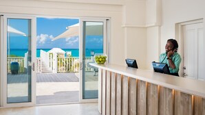 Un espace d’accueil avec vue sur l’océan, un comptoir de réception en bois et une porte en verre donnant sur une terrasse extérieure.