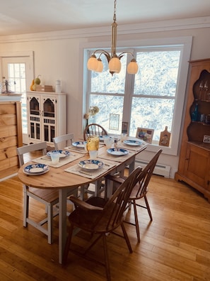 A dining room with a wooden table set for four, surrounded by chairs. A chandelier hangs above, and a window provides a view of a snowy landscape.