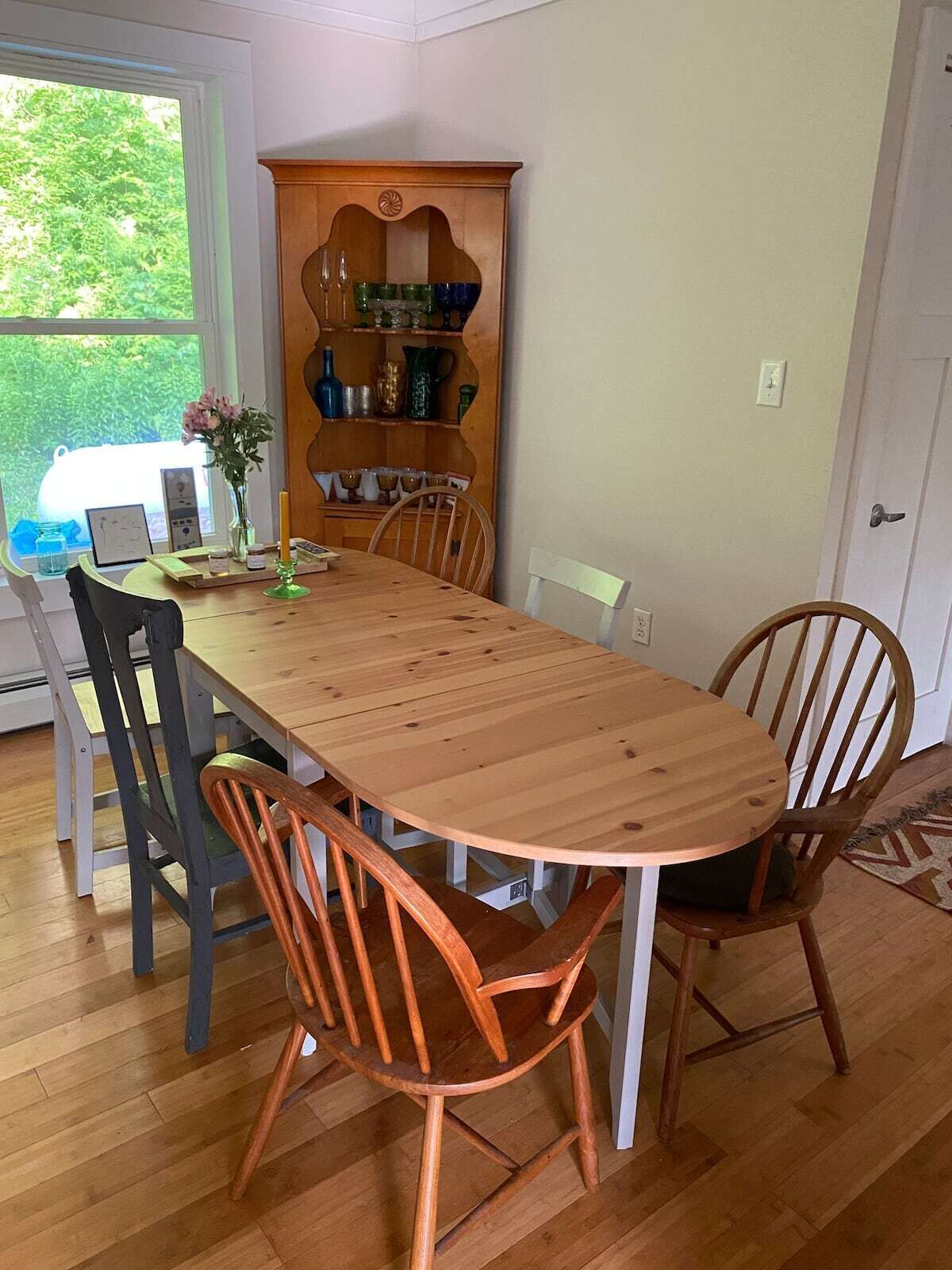 Un comedor con una mesa de madera y cuatro sillas, un armario de madera con vajilla, y un ventanal con vistas a zonas verdes.