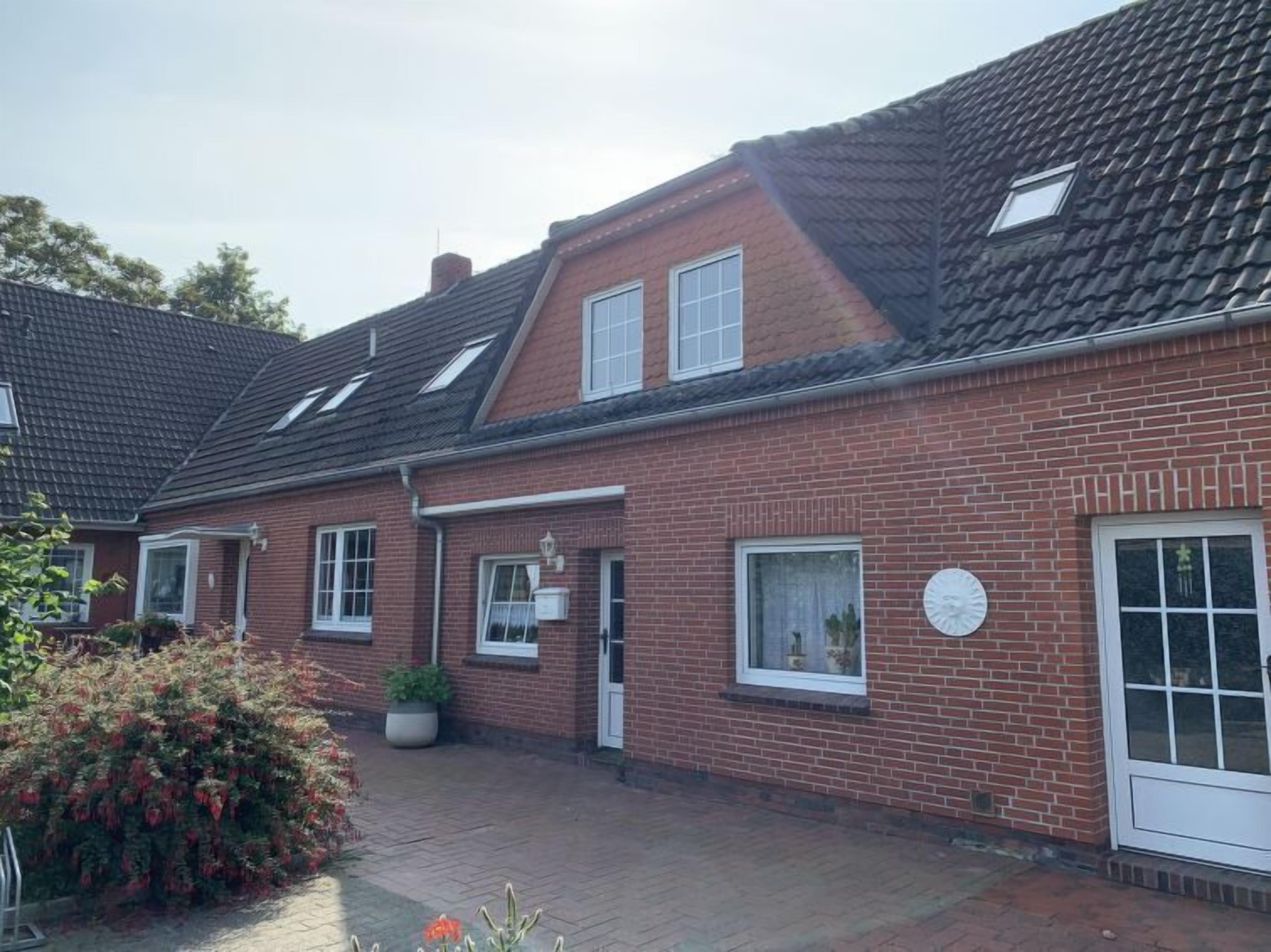 Une maison à deux étages, construite en briques, avec un toit en tuiles, un porche à l’avant et un petit jardin agrémenté de plantes en pot.