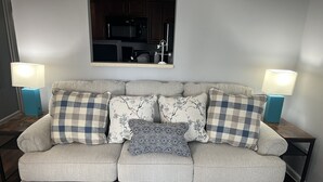 A grey sofa with patterned cushions, two side tables with lamps, and a kitchen visible in the background.