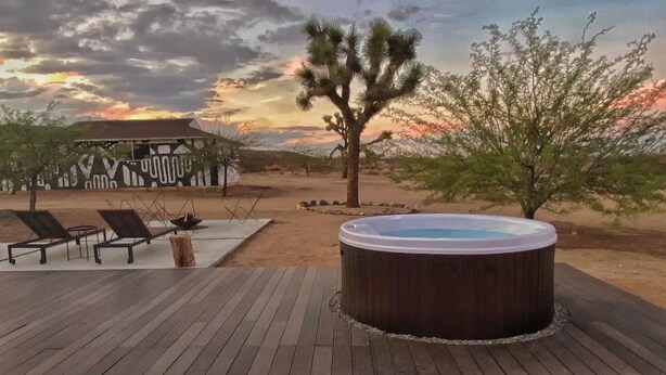 A hot tub on a wooden deck with lounge chairs, a desert landscape, and a building with a unique facade in the background.