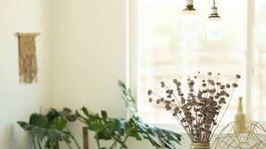 A dining area with a wooden table, two chairs, a vase with dried flowers, and hanging pendant lights.