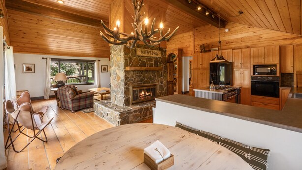 A cozy cabin interior with a stone fireplace, wooden ceiling, and a kitchen area. - Beautiful Log Farmhouse in Paradise Valley, Montana (Livingston)