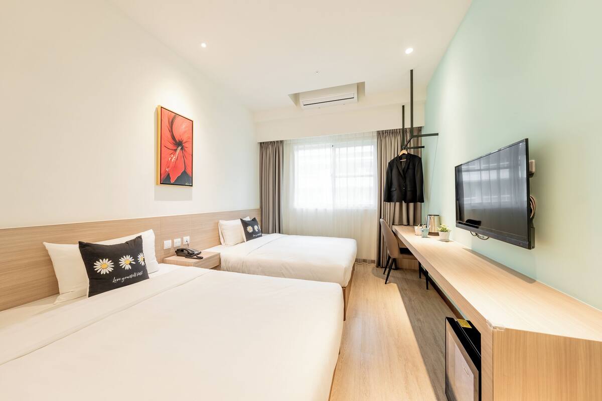 A hotel room with two beds, a TV, a desk, and a window with curtains.
