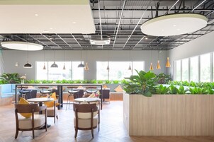 Un espacio interior moderno con suelo de madera, mesas y sillas redondas, grandes ventanales y plantas de interior.