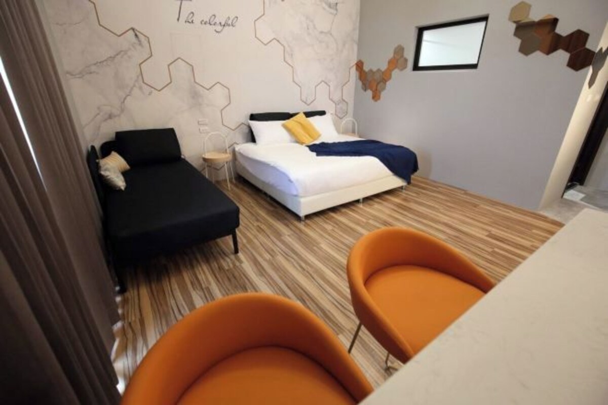 A modern hotel room with a bed, a sofa, and two orange chairs.