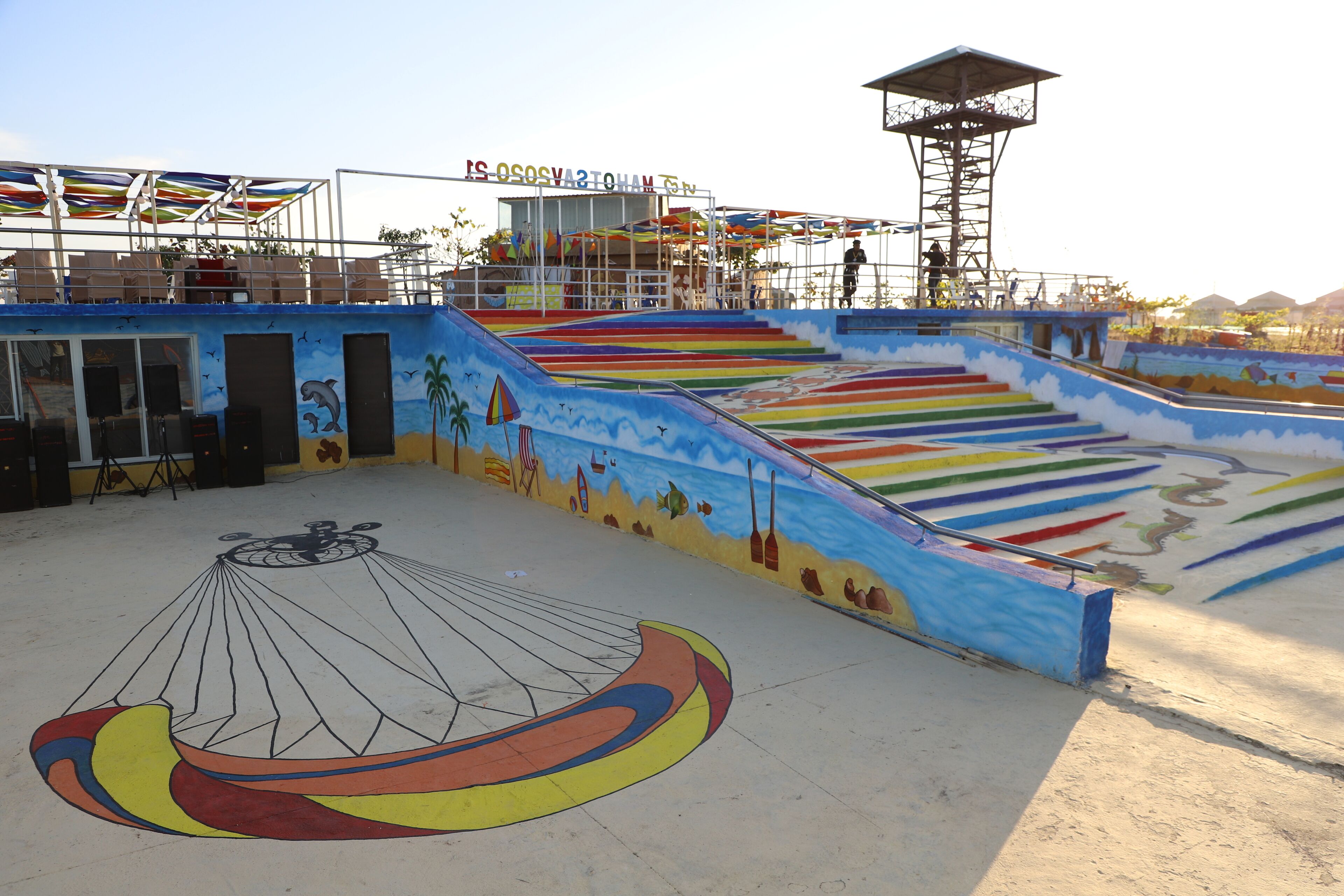 A colorful outdoor area with a mural of a sailboat, a stage, and a viewing platform.