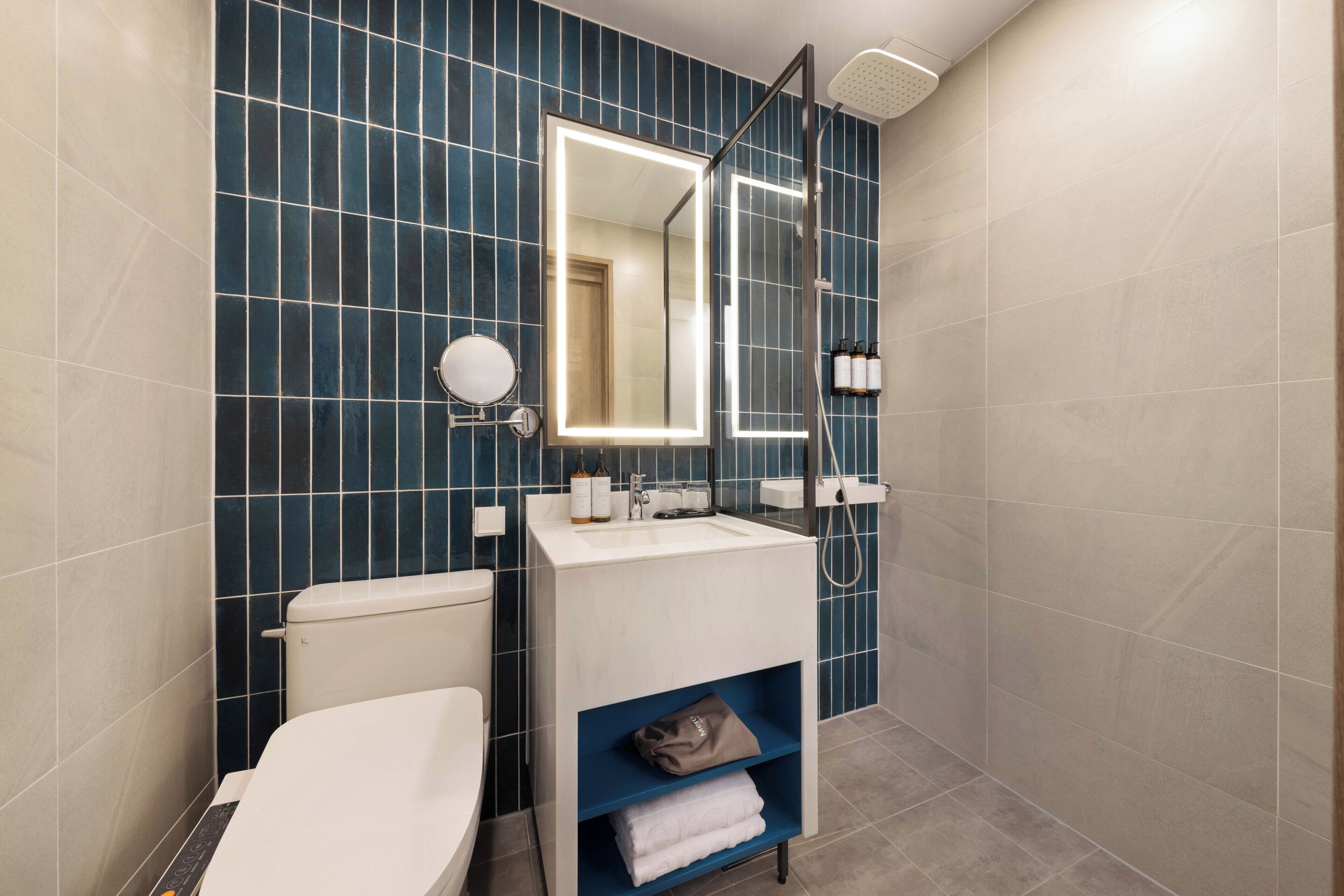 Un baño moderno con inodoro blanco, lavabo con espejo y ducha. Hay artículos de aseo en el lavabo y un estante para toallas.