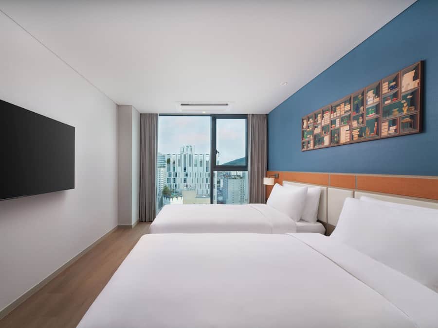 A hotel room with two beds, a large window, a flat-screen TV, and a wall-mounted artwork.