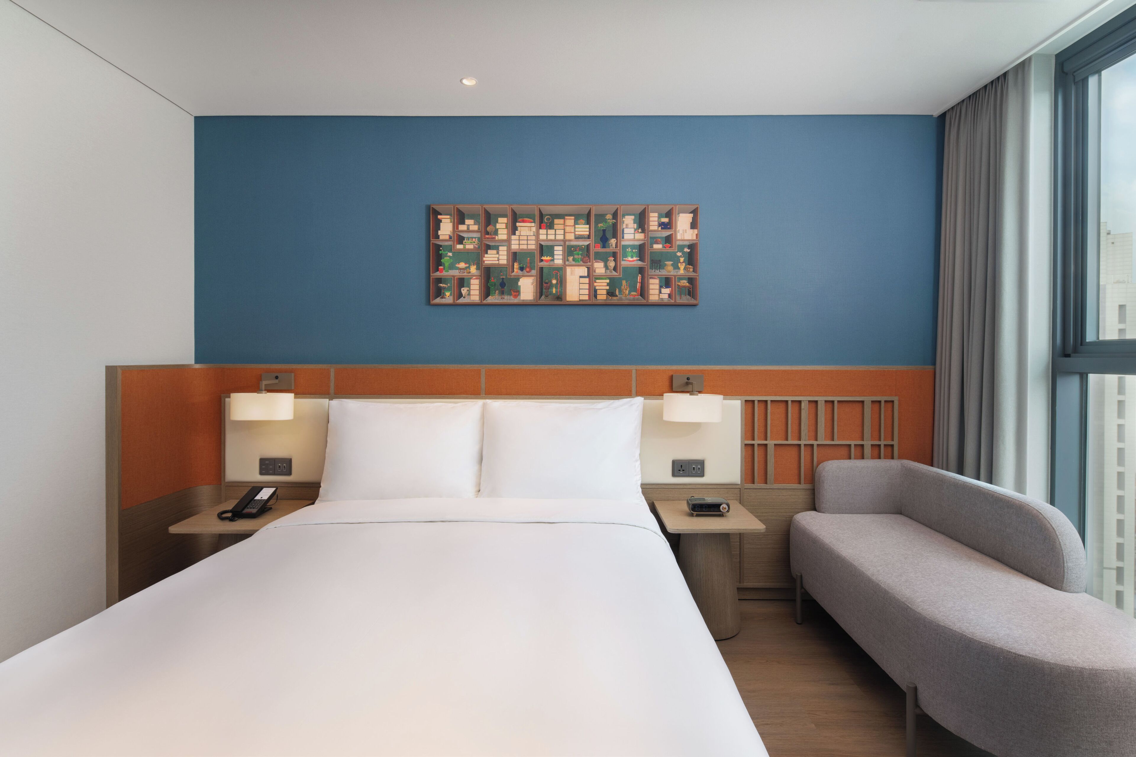 Ein Hotelzimmer mit einem großen Bett, zwei Nachttischen mit Lampen, einem grauen Sessel und einem an der Wand befestigten Kunstwerk.