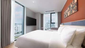 A hotel room with a large bed, a flat-screen TV mounted on the wall, and a city view through the windows.