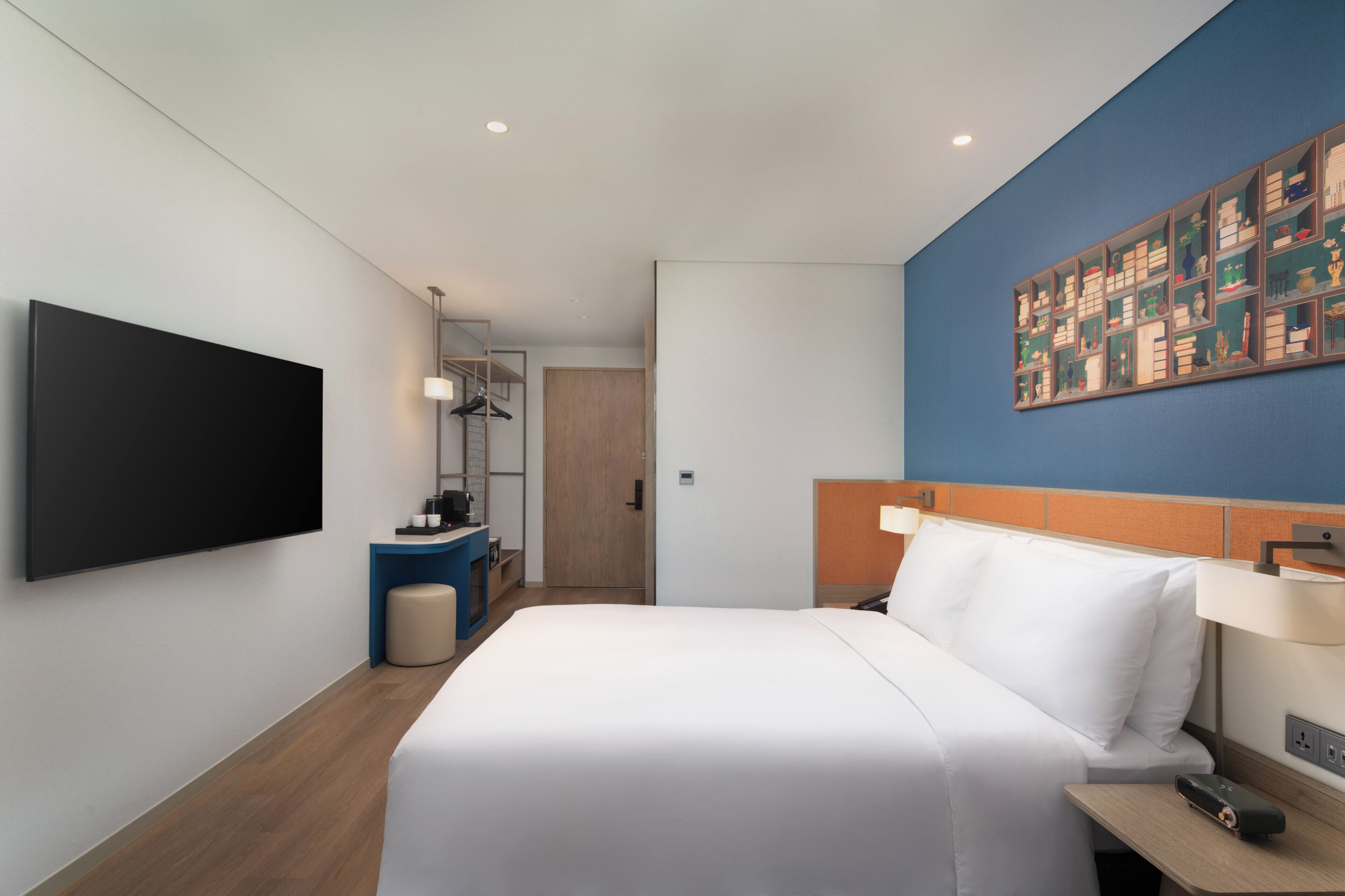 Une chambre d’hôtel moderne dotée d’un grand lit, d’un téléviseur à écran plat, d’un bureau avec une chaise et d’une étagère murale agrémentée d’objets décoratifs.