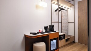 A modern hotel room with a wooden desk, a coffee machine, a mini-fridge, and a hanging coat rack.