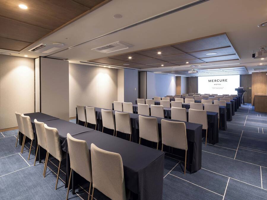 Een congreszaal met rijen tafels en stoelen, een groot scherm waarop tekst wordt getoond, en een houten plafond met inbouwverlichting.