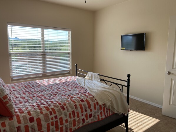 A bedroom with a bed, a window with blinds, a television on the wall, and a door. - Casita del sol (Tubac)