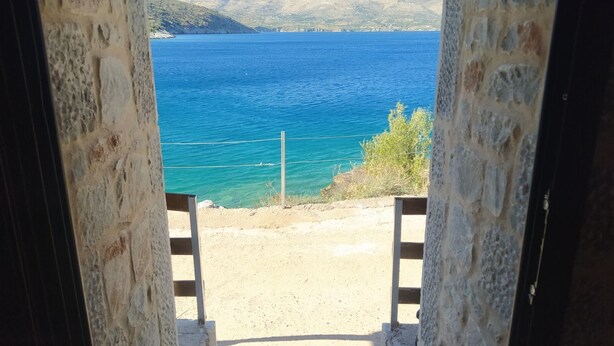 Une vue à travers un portail en pierre donnant sur une plage aux eaux d’un bleu limpide et sur des montagnes lointaines.