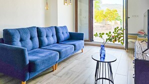 A modern living room with a blue sofa, a small round table with a vase, and a large sliding glass door leading to an outdoor area with a view of trees and a building. - Holiday Home "Cortijo Airen" with Pool, Mountain View & Wi-Fi (Cómpeta)