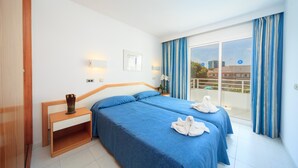 A hotel room with two beds, a wooden nightstand, a small table, a window with curtains, and a view of buildings outside.