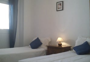 Une chambre d’hôtel avec deux lits, une table de chevet avec une lampe, un tableau encadré au mur et une fenêtre avec des rideaux.