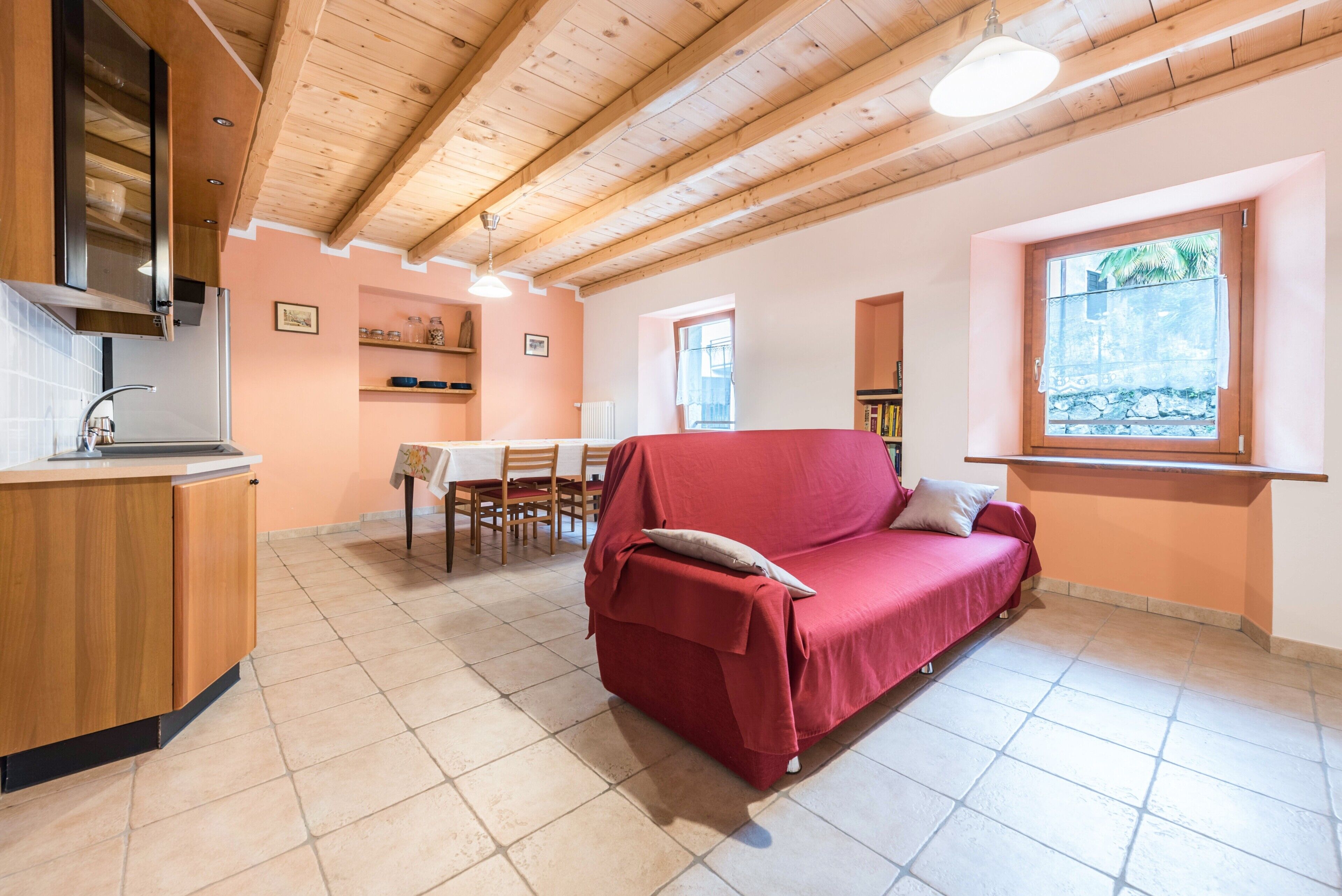A cozy living room with a red sofa, wooden ceiling beams, a tiled floor, and a window with a view.