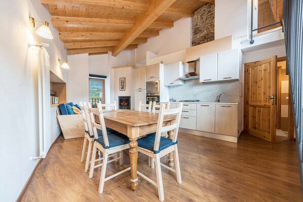 A wooden dining table with chairs, a kitchen with white cabinets, a stone wall, and a wooden ceiling.