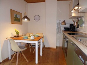 Uma área de refeições com uma mesa de madeira, cadeiras e um vaso de flores. Há um relógio na parede e uma cozinha com armários e eletrodomésticos.